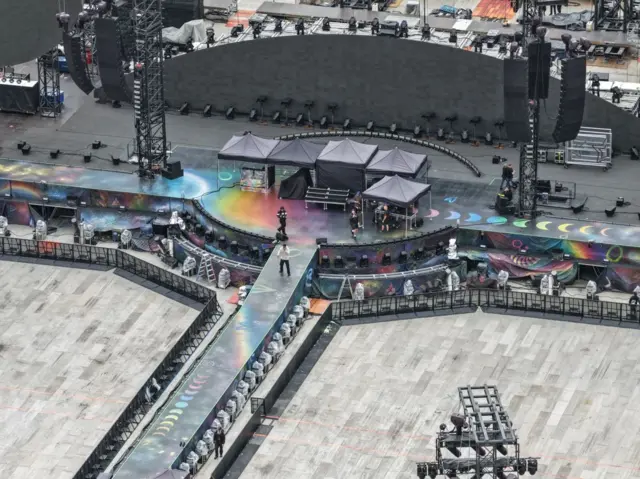 A large stadium's stage can be seen with several stalls on it with black canopies on the top. It's an aerial image. A man can be seen stood on the walkway just in front of the stage with a microphone. No one is in the standing area.