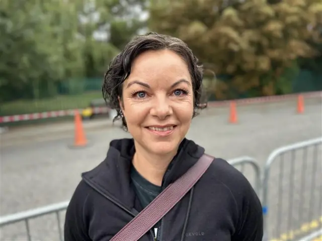 A head and shoulders shot of a woman with short brown hair smiling into the camera. She is wearing a black fleece and top with a purple strap across her shoulder. The background is blurred with metal railings and cones.