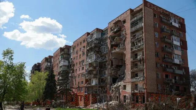 A red brick block of flats with severe damage along the front from where a strike hit, the sky is bright blue and there are trees in front of the buidling