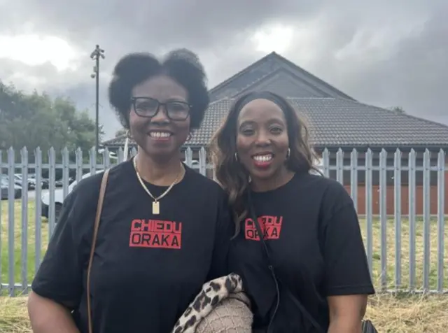 A head and shoulder shot of two women - Justina Oraka and Nkolika Ohaegb - smiling at the camera. They are stood outside and are wearing black t-shirts with red writing on reading "Chiedu Oraka".The woman on the left has short black hair and is wearing black-rimmed glasses. The woman on the right has long dark brown hair.