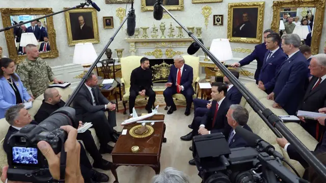 Wide shot of the inside of the Oval Office. Volodymyr Zelensky and Donald Trump are in the centre sitting on matching yellow brocade fabric chairs, their aides sitting to their sides on matching pale yellow couches. Overhead are four boom mics