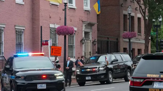 A black car bearing two flags - a US flag and the union jack - is seen in a street lined with brown stone buildings
