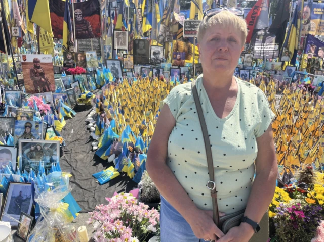 Natalia standing in Maidan Square, there are many flags, pictures of and tributes to killed Ukrainian soldiers behind her