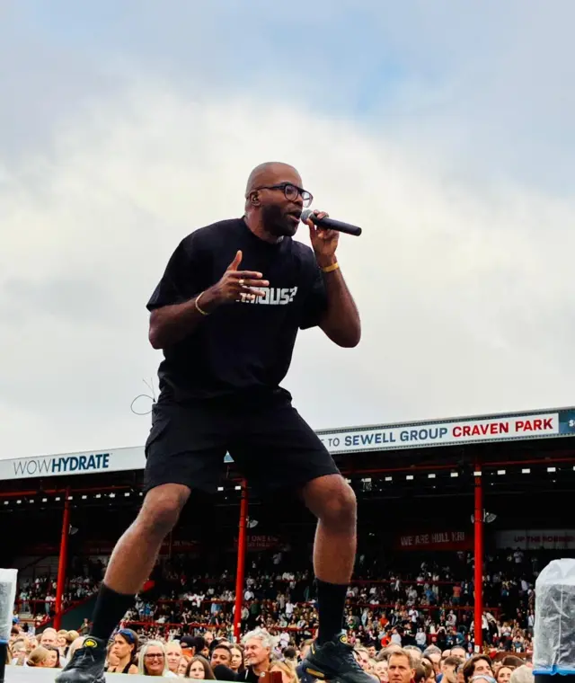 Chiedu Oraka on stage holding a microphone to his mouth. He is wearing a black t-shirt with white writing on, black shorts, black socks and black trainers. Crowds can be seen behind him.