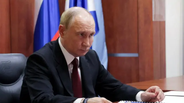 Putin sits behind a desk, wearing a suit and looks ahead.