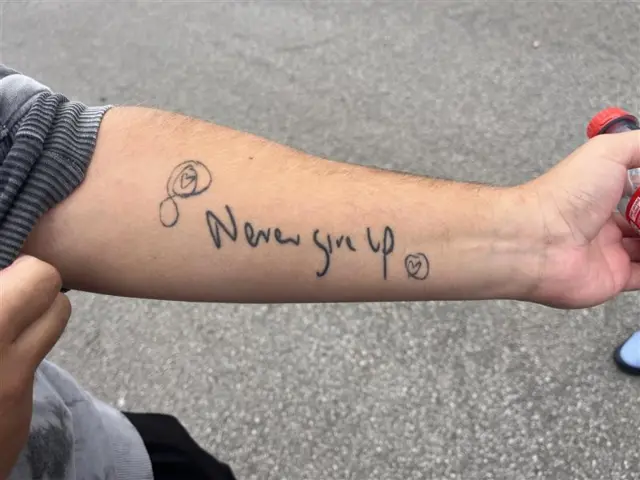 A man's arm with a tattoo of handwriting reading "Never Give Up" with an infinity sign with a love heart in a circle on the left side of the writing and a circled love heart on the right of the writing.