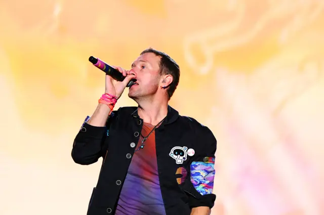 Coldplay frontman Chris Martin on stage holding a microphone to his mouth. He is wearing a black jacket and a multi-coloured t-shirt underneath.