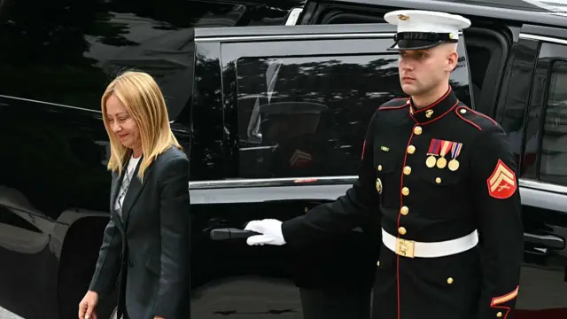 Giorgia Meloni in dark suit steps off a black car, a US marine holding the door open for her