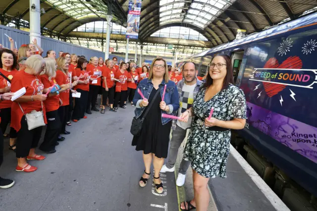Two women holding pin glowsticks stood either side of a man on a train platform. One woman has shoulder-length blonde hair and black glasses and is wearing a black dress, a denim jacket, a black bag and black sandals. The other woman has shoulder-length red hair and black glasses and is wearing a black and blue floral dress with black sandals. The man is wearing a grey checked shirt, a white t-shirt with a grey and yellow pattern on, grey trousers and white trainers. A group of people in black trousers and red t-shirts stand in a line to the left. There is a purple train on the right with a colourful design reading "SPEED OF SOUND" in capital letters.