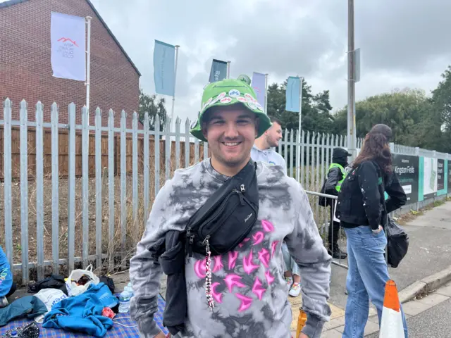 A head and shoulders photo of a man - Matty - smiling at the camera. He is wearing a green bucket hat with white badges on. He is wearing a grey tie-dye jumper, with a neon pink design on the front. He is also wearing a black bumbag around his chest.