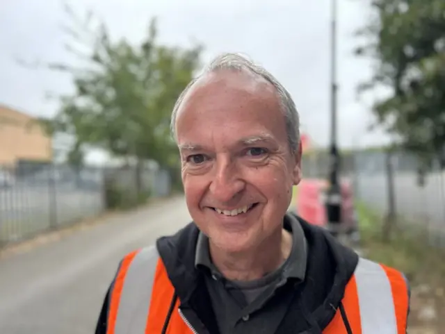 A head and shoulders shot of a man with short grey hair and a high vis jacket smiling into the camera. Behind him is a blurred path with railings each side.