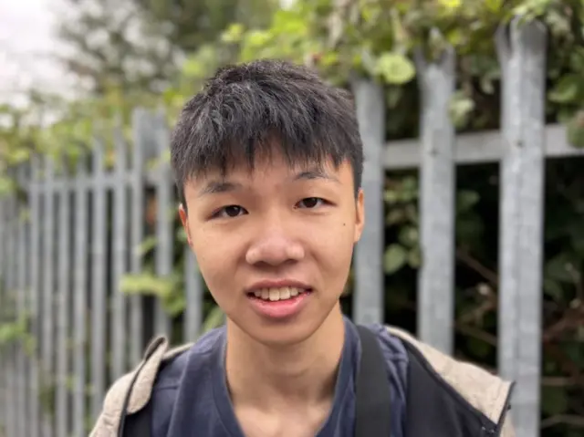 A young Chinese man with short black hair and a full fringe is smiling into the camera. He is wearing a navy top and jacket with blurred railings behind him.