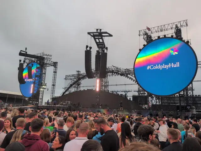 A stage with two large circular screens at either side. On the screen is a blue background, a rainbow and writing that reads "#ColdplayHull". Crowds can be seen looking towards the stage