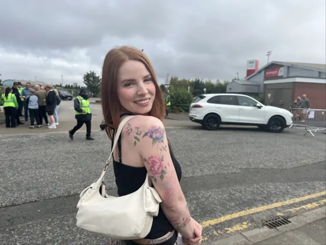 A photo of a woman - Angel - looking over her shoulder smiling at the camera. She is wearing a black vest and has a cream bag on her shoulder. She has colourful tattoos covering her right arm. People and cars can be seen in the background.