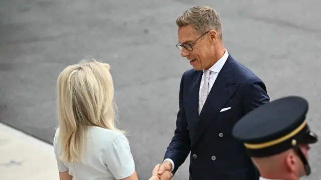 Stubb wears a double breasted navy suit and pale cream tie, he shakes the hand of a blonde woman in a white dress