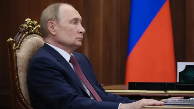 A side view of Putin sat at a table with his hands grasping the table in front of a Russian flag