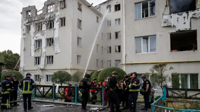 A badly damaged apartment block, windows are blown out  and part of it has collapsed. Firefighters are spraying water into one window to extinguish a fire