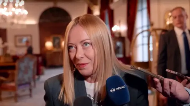 Giorgia Meloni speaking into a microphone in a room with a chandelier.