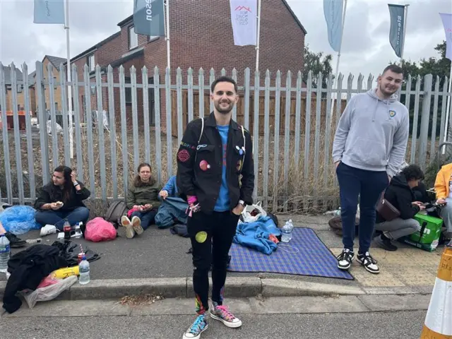 A man - Martin - standing outside, with his hands in his pockets, in front of a path, grey railings are behind him. He has short black hair and black facial hair. He is wearing black trousers and a black jacket with patches of planets on. He is also wearing colourful shoes with blue and pink laces and a blue t-shirt.