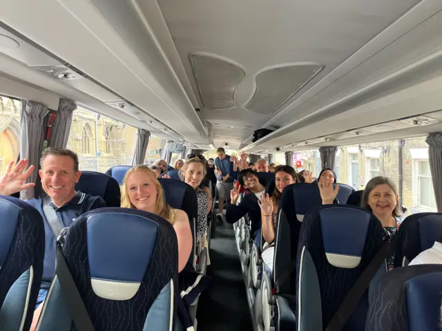 A photo taken from inside a bus. Passengers fill some of the seats and are smiling or waving at the camera. The seats are navy blue.