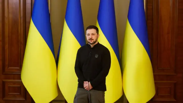 Zelensky wears a black top and stands in front of four Ukrainian flags.