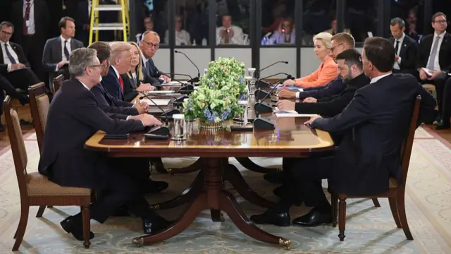 Wide shot of the leaders sitting at a wooden table in wooden chairs. On the left side are Keir Starmer, Emmanuel Macron, Donald Trump, Giorgia Meloni and Friedrich Merz. On the right side are Mark Rutte, Volodymyr Zelensky, Alexander Stubb and Ursula von der Leyen. In the background, inside white and grey plastic cubicles are the leaders' interpreters