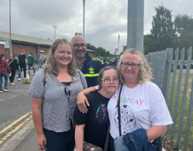 A family of four - a mother, her two daughters and her husband - stand in a group and smile at the camera