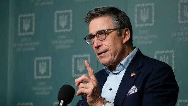 Anders Fogh Rasmussen raises his hand while speaking into a microphone.