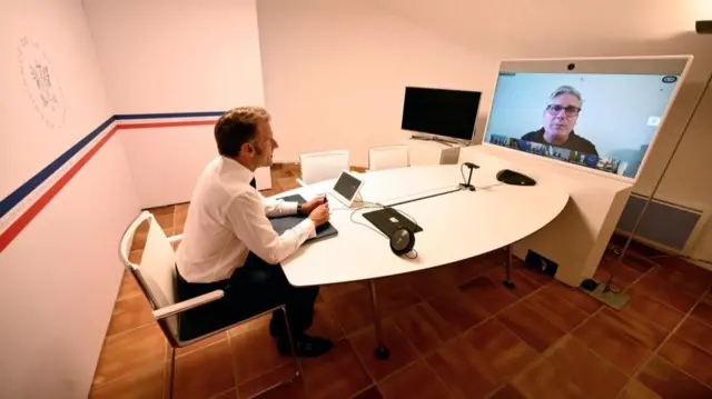 French President Emmanuel Macron sits at a desk in an office room looking at a large screen with UK Prime Minister Sir Keir Starmer speaking during a meeting of the "coalition of the willing"