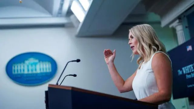 Karoline Leavitt pictured from the side standing at a podium in the White House briefing room, wearing a white dress and raising a hand in the air.