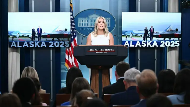 Karoline Leavitt stands at a podium giving a White House press briefing. Behind her are two screens with the words "Alaska 2025" and a picture of Vladimir Putin and Donald Trump meeting in the US state on Friday. The back of journalists' heads in the room listening to Leavitt are also pictured.