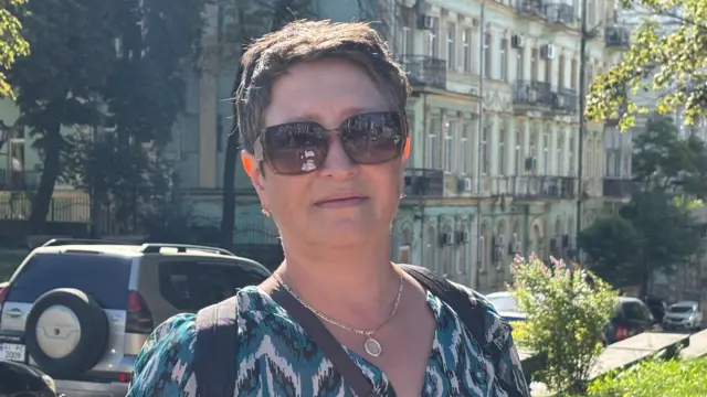 Wearing dark sunglasses Viktoria looks directly at the camera. Her backpack and handbag straps hang over her shoulder. Parked cars and Kyiv buildings are behind her in the background