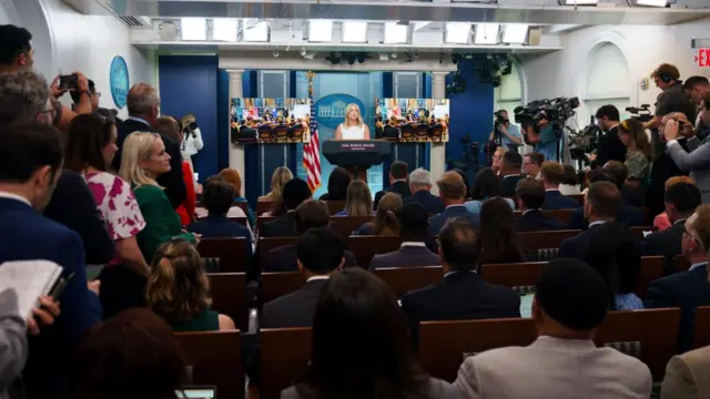 White House Press Secretary Karoline Leavitt, wearing a white dress and standing behind a podium, speaks to the media during a press briefing at the White House. Seated journalists and some standing are in the foreground of the photo.