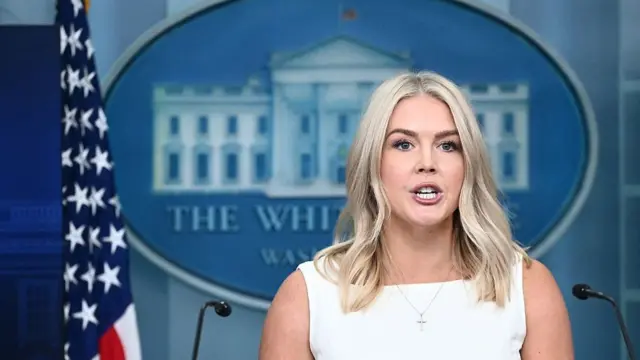 Karoline Leavitt speaks during a White House press briefing wearing a white dress. The White House logo and a US flag are behind her.