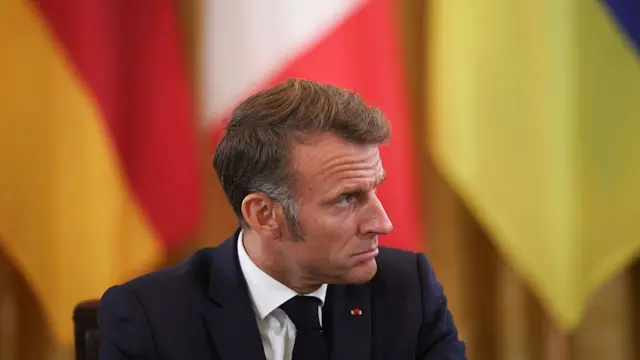 Emmanuel Macron looks right out of frame sitting in front of three flags.