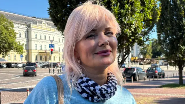 Veronyka looks directly at the camera. She is wearing a blue top with a handbag strap and a black and white scarf around her neck