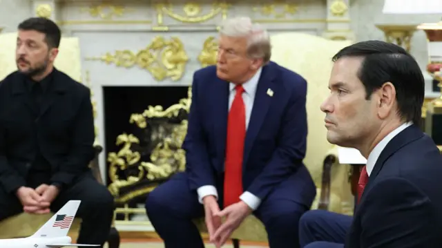 Side view of Marco Rubio in the white house wearing a suit. In the background, seated near him, are Zelensky and Trump, both wearing suits.