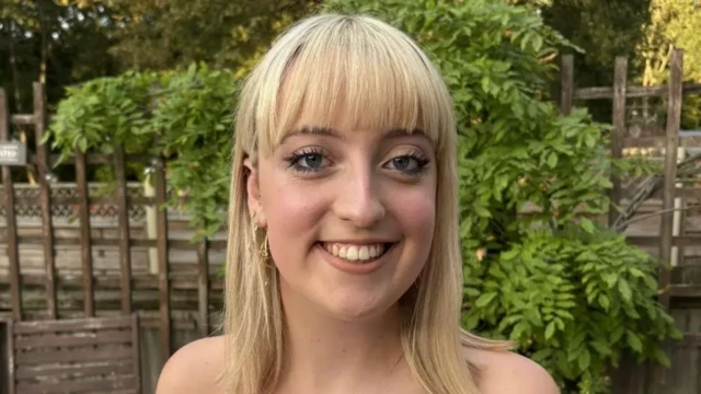 Imogen who has blonde mid length hair and a fringe looks at the camera smiling. Behind her is a brown garden fence and green leaves.