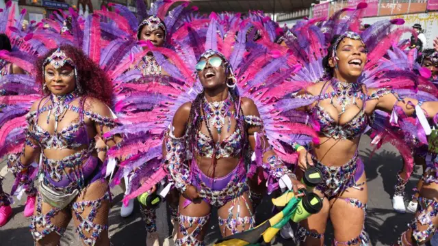 Four female dancers in purple bejewelled dancewear and pink and purple feathered carnival plumes dance on the street outside a tube station