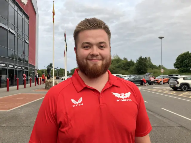 William Llewellyn Davies wearing a red scarlets shirt. He is standing outside Parc y Scarlets