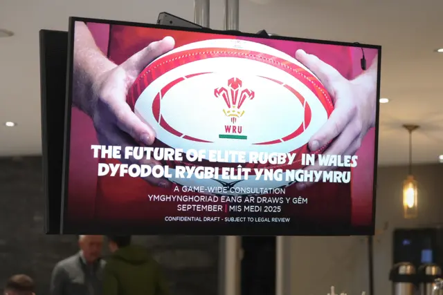 A large digital board shows a WRU rugby ball as the background with text saying 'The future of Elite Rugby in Wales'. People can be seen chatting in the background behind the screen.