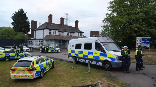 Several police cars and vans are parked around the Bell Hotel, which is an old building surrounded by a metal temporary fence.