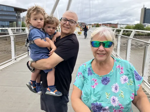 Denise and Jaff smiling looking at the camera standing on a bridge in Newport. On the right is Denise, who has white hair and is wearing a turquoise t-shirt and green mirrored sunglasses. On the left, is Jaff, who is holding two young twin boys. He has very short white hair and is wearing a black t-shirt and black glasses.
