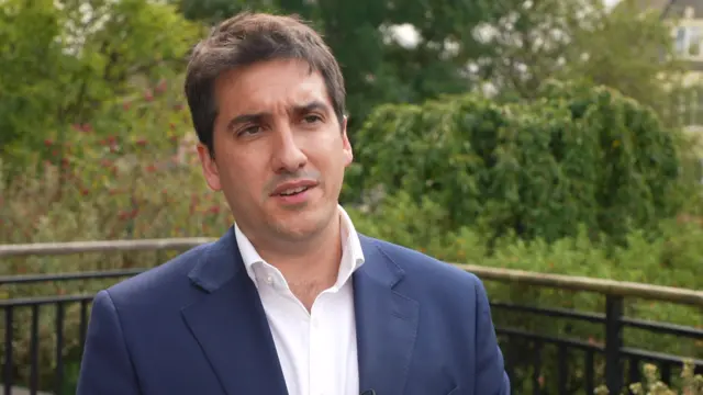 A man with brown hair in a blue blazer and white shirt stood outside trees