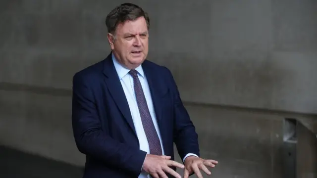 Mel Stride in a blue suit, and red tie, hands outstretched, brown hair.
