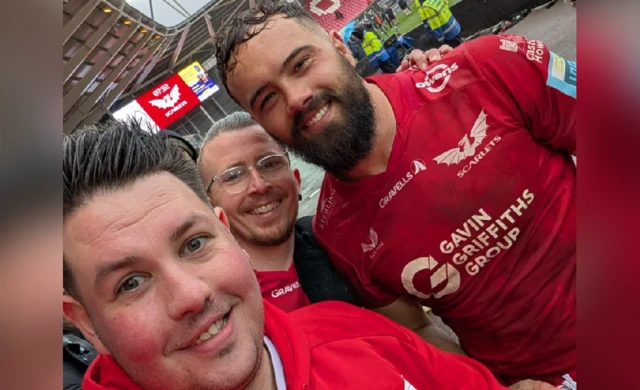 Selfie of Ben Griffiths smiling with a friend and Josh Mcleod after a Scarlets match. On the left, Ben wears a red Scarlets hoodie. He has short spikey brown hair. In the middle is his friend, who is wearing a red Scarlets t-shirt, clear glasses and has short grey hair. Josh, who is on the right, is wearing a red Scarlets kit. He has short brown hair and a beard.