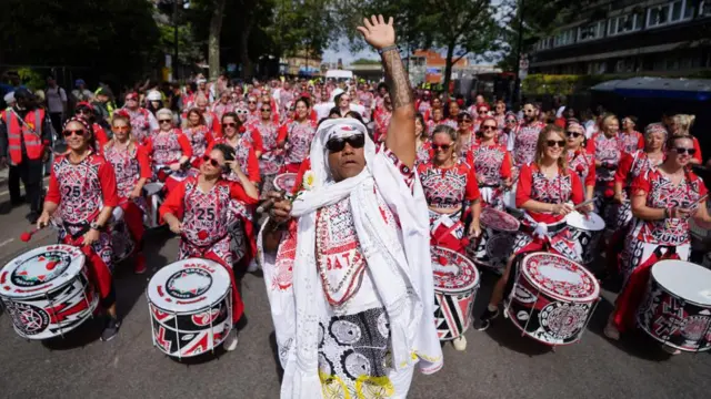 A large Brazilian drumming band  - wearing matching white, red and black t-shirts and trousers - passes through the streets in Westbourne Park during the adult's parade in 2024