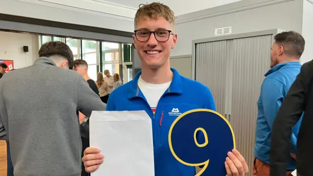 Kacper is stood in a crowded school hall holding his results envelope in one hand, and a large number nine in the other. He wears glasses and is smiling at the camera wearing a quarter-zip blue jumper