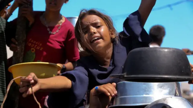 A young girl wearing a blue top holds a bucket out asking for aid, she is surrounded by other bowls and pots and people