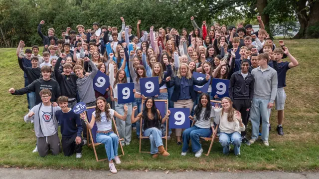 A group of pupils stand in a crowd - some with their arms in the air. Others sit on deckchairs and hold posters reading '9'